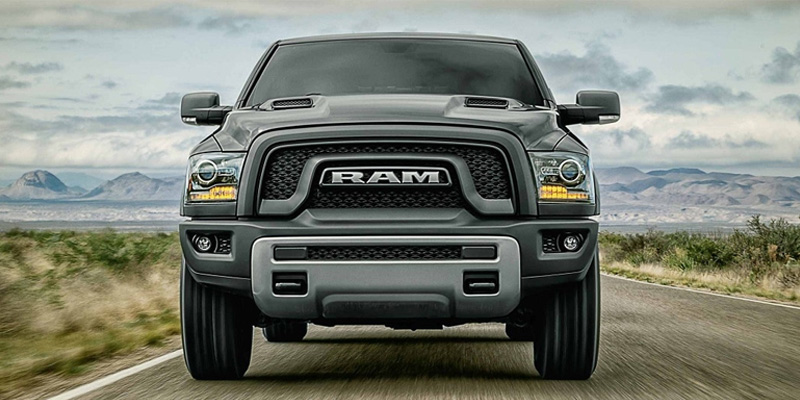 A dark gray RAM 1500 driving down a highway straight towards the camera, backed by mountains and a cloud-filled sky.
