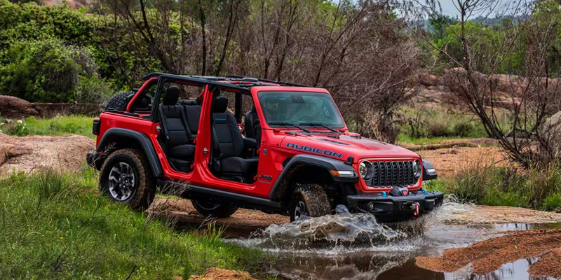 A red Jeep wrangler with the doors off, in a forested area, about to ford a creek.