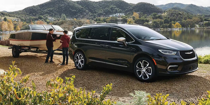 A black Chrysler Pacifica parked near a lake, with a family unloading a boat from the trailer, showcasing the vehicle's towing capability and family-friendly design.
